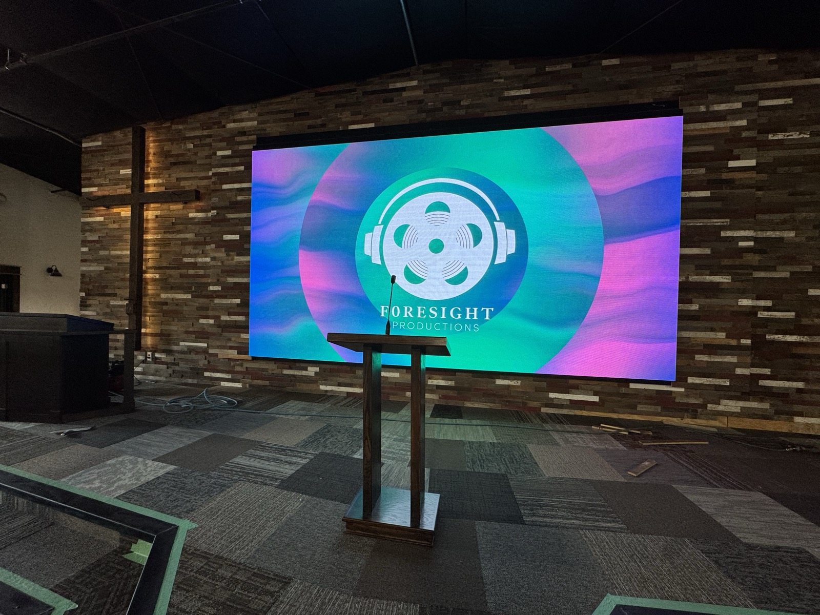Church stage LED video wall mounted on a wood-plank accent wall beside a wooden cross and pulpit